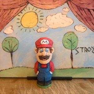 Può includere: Una statuetta di legno intagliata a mano di Mario, con un berretto rosso con una "M" bianca, camicia rossa e tuta blu. La statuetta è davanti a uno sfondo dipinto di un cielo soleggiato, nuvole, alberi e la parola "STROZYK".