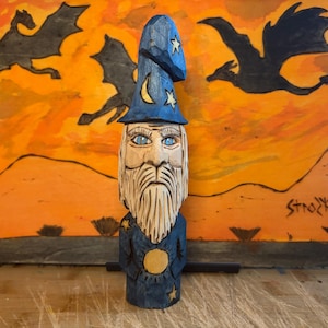 Può includere: Statuetta di mago in legno intagliato a mano con una lunga barba bianca, occhi blu e un cappello blu ornato di stelle e una luna crescente. Il mago indossa una veste blu con un emblema solare. Lo sfondo presenta draghi contro un cielo arancione.