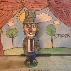 Può includere: Una statuetta di legno di un clown triste con un cappello marrone, pantaloni blu e una cravatta viola. Il clown è in piedi davanti a uno sfondo dipinto di un palco con tende rosse, un cielo blu e due alberi. Lo sfondo ha anche la parola "STROZK" scritta su di esso.