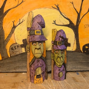 Può includere: Due figurine di strega intagliate nel legno con cappelli viola e scope. La strega più grande tiene un sacchetto di dolcetto o scherzetto. Le figurine sono posizionate su uno sfondo dipinto di un cimitero e una luna piena.