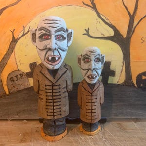 Può includere: Due figurine di vampiro in legno intagliate con occhi rossi e zanne. La figurina più grande indossa un cappotto marrone e la figurina più piccola indossa un cappotto marrone. Le figurine sono in piedi su una superficie di legno davanti a uno sfondo dipinto con una luna e alberi.