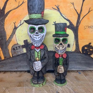 Può includere: Due figure in legno intagliate di scheletri che indossano cappelli a cilindro e abiti. Gli scheletri hanno volti dipinti di verde e bianco con occhi neri. Le figure sono in piedi su una superficie di legno davanti a uno sfondo dipinto con alberi e una luna.