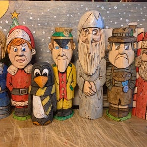Può includere: Una collezione di sette figure di legno intagliate. Le figure includono una donna in un vestito blu, una persona in un costume da Babbo Natale, un pinguino e altri personaggi in vari abiti. Le figure sono dipinte con colori vivaci e hanno tratti del viso dettagliati.