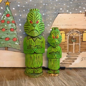Può includere: Due figure di legno verde del Grinch con le braccia incrociate davanti a una scena natalizia dipinta. Lo sfondo presenta un albero di Natale, una casa con la scritta "Santa's Workshop" e neve che cade. Le figure del Grinch hanno gli occhi rossi.