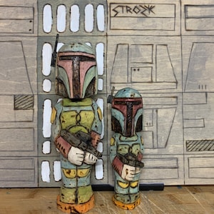 Può includere: Due sculture in legno di un personaggio con elmetto e armatura, che tiene in mano un blaster. Le sculture sono dipinte in tonalità di verde, blu, arancione e rosa. Lo sfondo è un pannello di legno grigio con la scritta "STROZK" in alto.