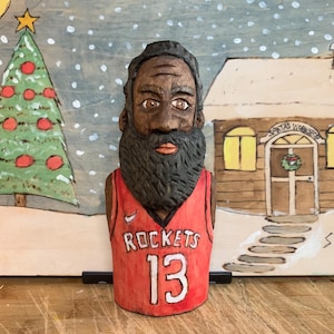 Può includere: Busto in legno intagliato di un uomo con una maglia rossa dei Rockets con il numero 13. Il busto è ambientato su uno sfondo dipinto di una scena natalizia con un albero, una casa e neve che cade.