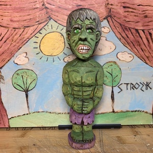 Può includere: Una statuetta di legno intagliata a mano di Hulk, dipinta di verde con pantaloncini viola. La statuetta è in piedi su un palco con uno sfondo dipinto di un cielo blu, nuvole, un sole e due alberi. Lo sfondo ha anche la parola "STROZK" scritta su di esso.