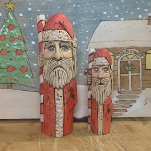 Può includere: Due figurine di Babbo Natale intagliate nel legno, una più grande dell'altra, entrambe con abiti di Babbo Natale rossi e bianchi e che tengono bastoncini di zucchero. Le figurine sono in piedi su una superficie di legno davanti a una scena invernale dipinta.