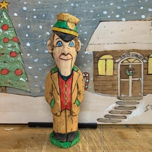 Può includere: Figura in legno intagliata a mano di un uomo in uniforme, con cappello verde e oro. Indossa un gilet rosso e una giacca marrone chiaro. Lo sfondo presenta un albero di Natale, una scena innevata e un edificio con "Santa's Workshop".