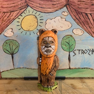 Può includere: Una statuetta in legno intagliata a mano di un personaggio di Star Wars, che indossa un costume arancione e marrone. La statuetta è in piedi su una base verde ed è posizionata su uno sfondo dipinto di un palco con tende, un sole, nuvole e alberi. Lo sfondo include anche la parola "STROZYK" scritta in un font stilizzato.