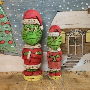 May include: Two wooden carvings of the Grinch, one larger than the other, both wearing Santa hats and red and white outfits. The carvings are standing on a wooden surface in front of a painted backdrop of a snowy scene with a Christmas tree and a house.