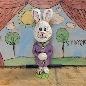 Può includere: Una statuetta di coniglio in legno intagliata a mano che indossa un cappotto viola. Il coniglio ha la pelliccia bianca, gli occhi neri e un naso rosa. La statuetta è in piedi su una base di legno con uno sfondo verde. Lo sfondo è una scena dipinta con un sole, nuvole e alberi.