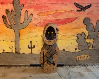 Puede incluir: Una figurita de madera de un personaje de Star Wars, con una túnica marrón con capucha y sosteniendo un arma. La figurita está de pie frente a un fondo de madera pintado que representa un paisaje desértico con cactus y una puesta de sol.