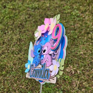 May include: A blue and pink cake topper featuring two cartoon characters, Stitch and Angel, with a pink number 9 and the name "Camila" on a blue and white background.