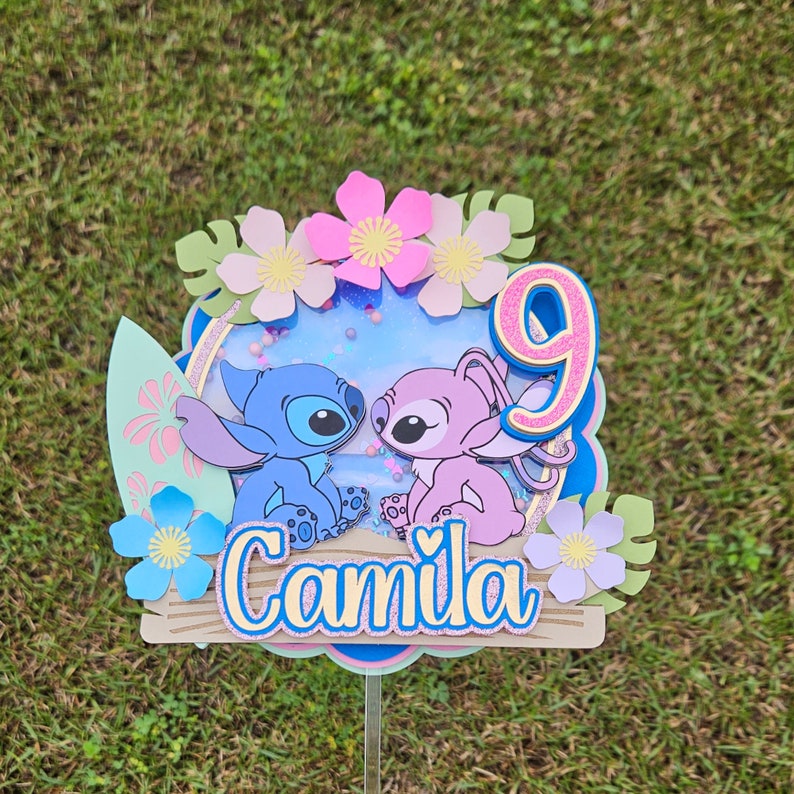 May include: A blue and pink Stitch and Angel cake topper with a pink number 9 and the name "Camila" on a wooden base. The topper is surrounded by flowers and leaves.