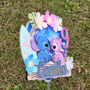 May include: A blue and pink cake topper featuring Stitch and Angel from the Disney movie Lilo & Stitch. The topper is decorated with flowers, glitter, and the name "Camila".