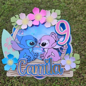 May include: A blue and pink Stitch and Angel cake topper with a glitter background and a surfboard. The topper also includes a pink number 9 and the name "Camila" in gold letters.