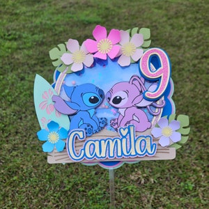 May include: A blue and purple Stitch and Angel cake topper with a pink number 9 and the name "Camila" on a wooden sign. The topper is decorated with flowers and leaves.