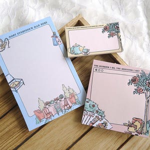 Op de afbeelding: Drie notitieblokken met het thema Alice in Wonderland. Het grootste notitieblok heeft de tekst "MOST EVERYONE'S MAD HERE" en illustraties van een klok, een fles en een tafelsetting. De andere notitieblokken hebben illustraties van een theeservies, rozen en speelkaarten.