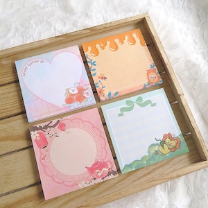 May include: Four square memo pads with unique designs. One pad has a heart shape with the text "never lychee go" and a cartoon lychee design. Other pads feature honey, a lion, and a pink floral design. The pads are in a wooden tray.