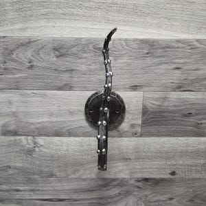 May include: A handcrafted, wrought iron wall hook. The hook features a circular mounting plate and a decorative branch-like design with small, raised accents. The metal has a dark, textured finish, and the hook is mounted on a wooden background.