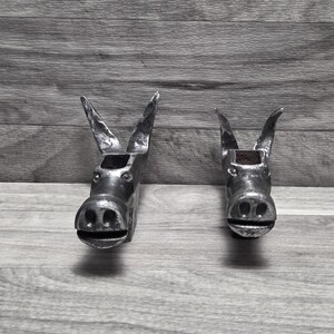 May include: Two metallic donkey head sculptures crafted from dark, textured iron. Each figure features prominent upright ears, a rounded snout with visible nostrils, and a horizontal mouth line, showcasing a raw, industrial finish with visible metalwork.