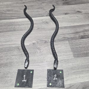 May include: Two dark metal snake sculptures with textured bodies and angled heads. Each snake is positioned above a small, dark square with a number and two green dots. The squares are labeled "1" and "2".