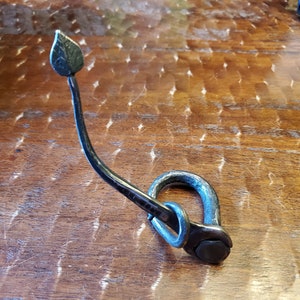 Puede incluir: Un gancho de metal negro con un diseño de hoja en la parte superior. El gancho está doblado en un bucle en la parte inferior y tiene una tuerca y un tornillo de fijación.