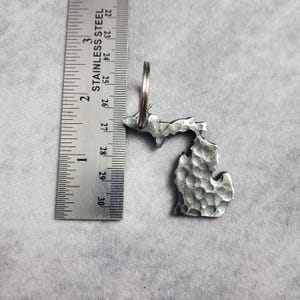 Puede incluir: Un llavero de metal plateado con forma del estado de Michigan. El llavero tiene una textura con un acabado martillado y tiene un anillo para llaves.