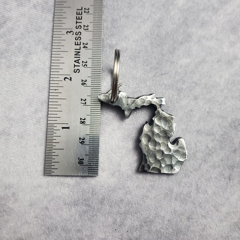 Puede incluir: Un llavero de metal plateado con forma del estado de Michigan. El llavero tiene una textura con un acabado martillado y tiene un anillo para llaves.