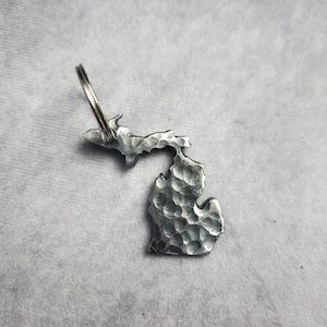 Puede incluir: Un llavero de metal plateado con forma del estado de Michigan. El llavero tiene una textura martillada y un anillo de llave plateado.