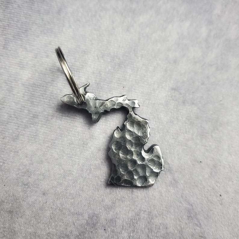 Puede incluir: Un llavero de metal plateado con forma del estado de Michigan. El llavero tiene una textura martillada y un anillo de llave plateado.