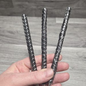 May include: Three metal pens with a textured, spiral design. The pens are held in a hand, showcasing their pointed tips and the detailed craftsmanship of the metalwork. The pens have a dark, metallic finish.