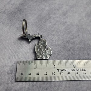 Puede incluir: Un llavero de metal plateado con forma del estado de Michigan. El llavero mide aproximadamente 5 cm de alto y tiene una textura martillada. Est&aacute; unido a un anillo de metal plateado.