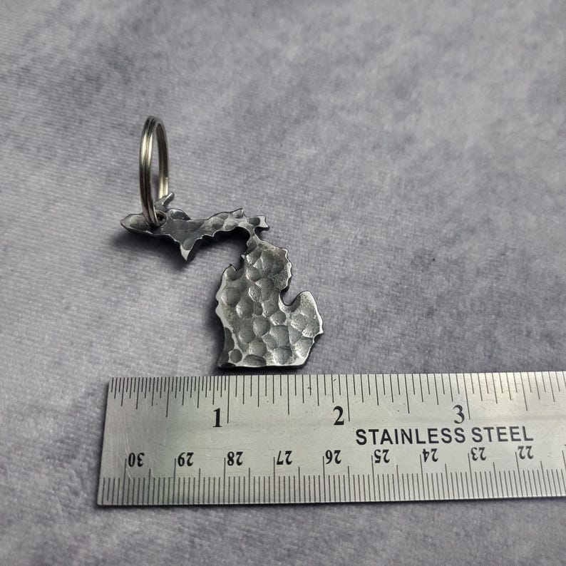 Puede incluir: Un llavero de metal plateado con forma del estado de Michigan. El llavero mide aproximadamente 5 cm de alto y tiene una textura martillada. Est&aacute; unido a un anillo de metal plateado.