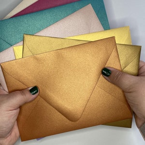 May include: A stack of colorful envelopes in various shades of gold, green, pink, and beige. The envelopes are made of textured paper and have a metallic sheen.