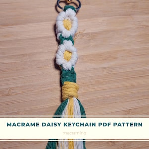 May include: A macrame daisy keychain with a gold ring. The keychain is made with green, white, and yellow yarn. The pattern is for a PDF download. The text "MACRAME DAISY KEYCHAIN PDF PATTERN" and "macraming" are visible.