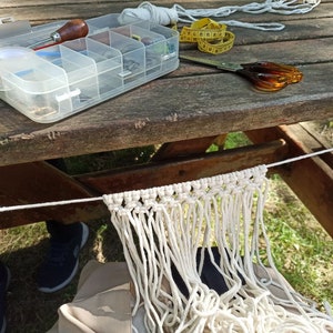 Puede incluir: Un tapiz de macramé blanco con flecos, colgado de una cuerda blanca atada a una mesa de madera. Una caja de plástico transparente con varias herramientas y una cinta métrica amarilla están sobre la mesa.