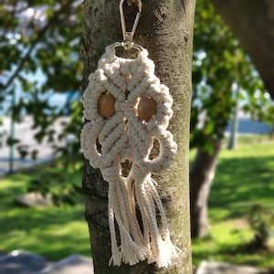 May include: A white macrame keychain with two wooden beads and a silver metal clasp. The keychain is hanging from a tree branch.