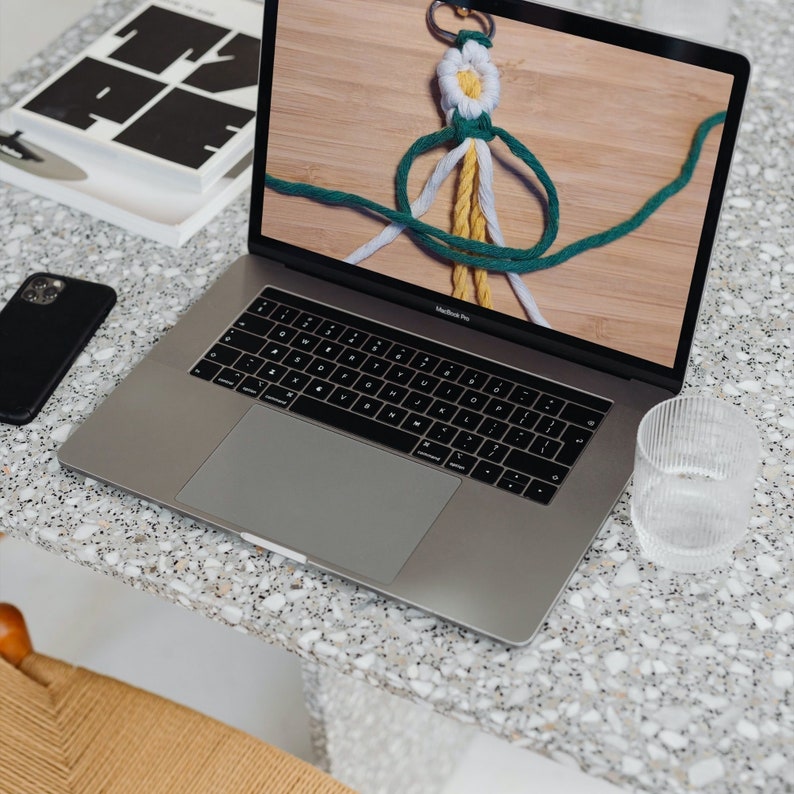 May include: A silver laptop computer with a white and green string knot displayed on the screen. The laptop is on a light gray speckled surface.