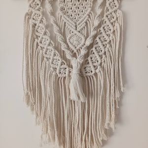 以下が含まれることがあります： 幾何学模様とフリンジが付いた白いマクラメの壁掛け。木製の棒に吊り下げられています。