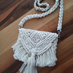 Peut inclure: Un sac en crochet blanc avec une bordure en franges et un pompon. Le sac a une longue bandoulière et est fait de macramé.