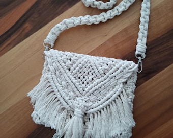 May include: A white crocheted bag with a fringe trim and a tassel. The bag has a long strap and is made of macrame.