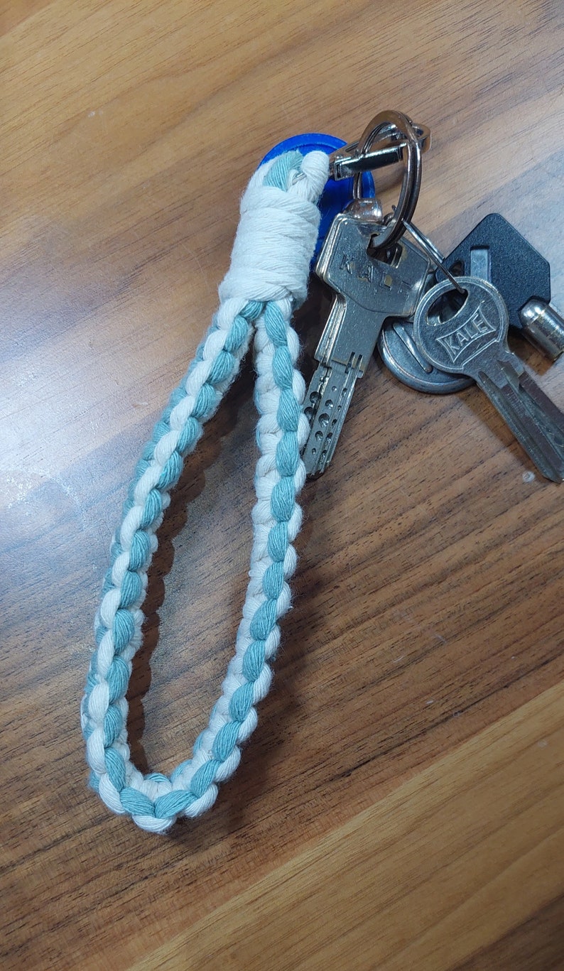 May include: A white and light blue braided rope keychain with a silver key ring and keys attached. The keys have silver metal bodies and black plastic handles.