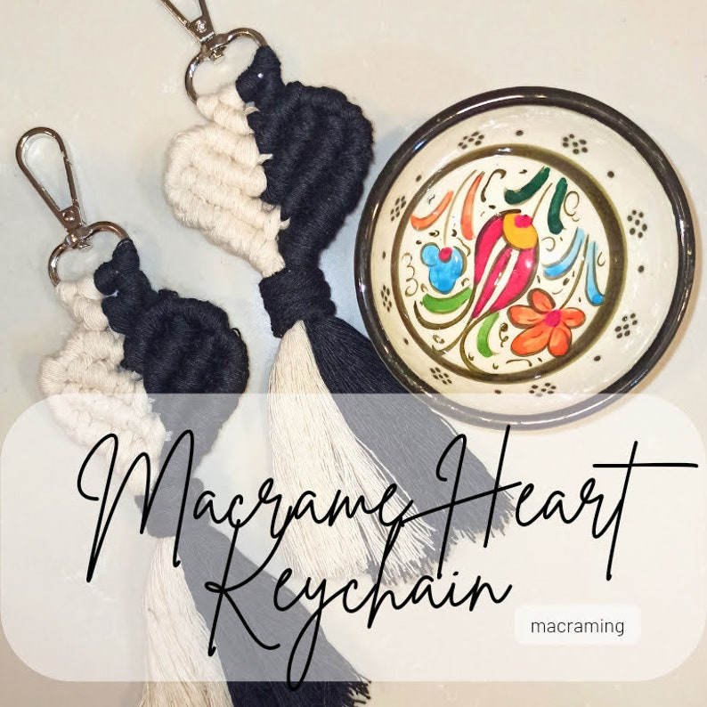 May include: Two macrame heart keychains with black and white yarn. The keychains have tassels and are hanging from silver key rings. The keychains are on a white surface with a small decorative bowl.
