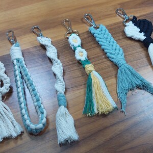 May include: A collection of six handmade macrame keychains with various designs and colors. The keychains feature a variety of patterns, including a skull, a flower, and a braid. The keychains are made with natural cotton cord and have a tassel at the bottom.