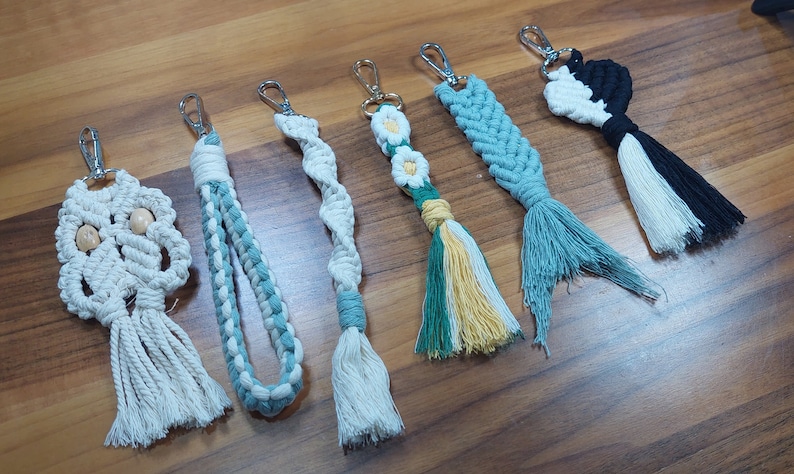 May include: A collection of six handmade macrame keychains with various designs and colors. The keychains feature a variety of patterns, including a skull, a flower, and a braid. The keychains are made with natural cotton cord and have a tassel at the bottom.