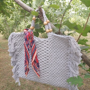 Puede incluir: Un bolso tote de crochet gris con detalles de flecos y asas de cuentas de madera. Una bufanda estampada en rojo, azul y blanco está metida en el bolso.
