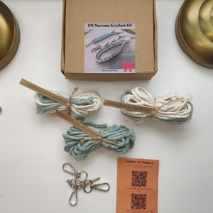 May include: DIY macrame keychain kit with two balls of white and blue cord, two balls of blue cord, four silver key rings, and a card with a QR code for a video tutorial. The box is labelled "DIY Macrame Keychain Kit".