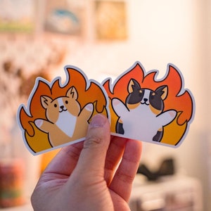 May include: Two colorful stickers featuring cartoon dogs in flames. The first sticker shows a corgi with orange and white fur, wearing a red collar. The second sticker shows a black and white cat with a red collar.